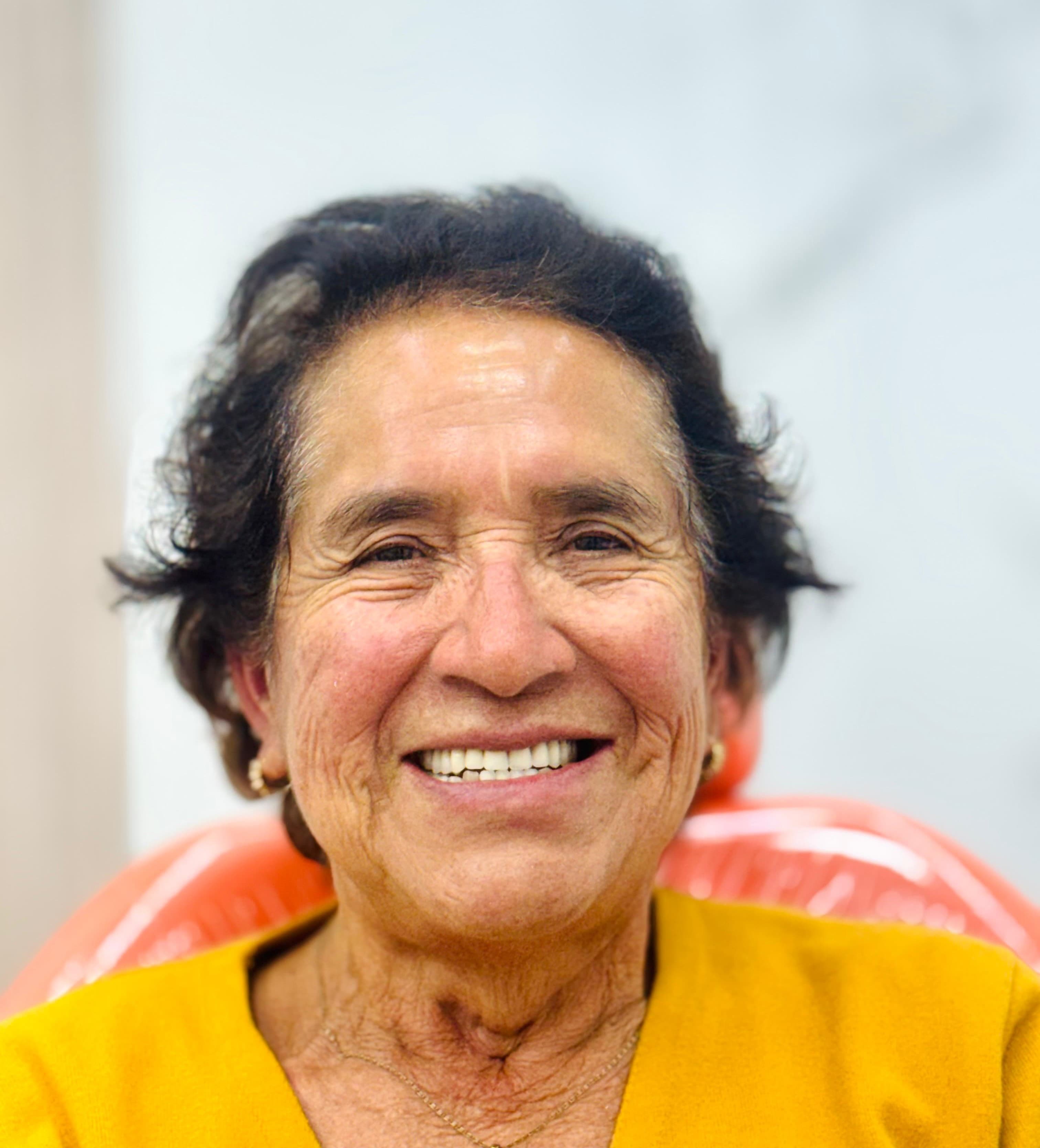 Smiling elderly woman with dark wavy hair wearing a yellow top and gold hoop earrings.