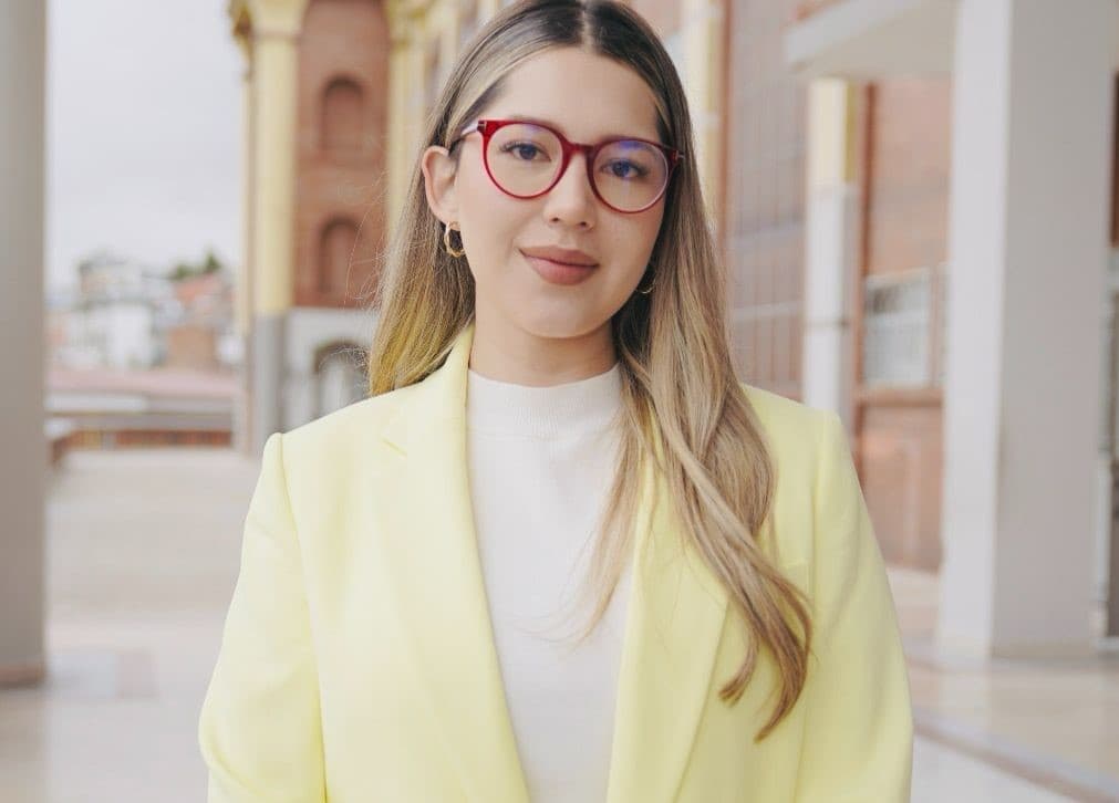Woman with long blonde hair wearing red glasses and a pale yellow blazer outdoors.