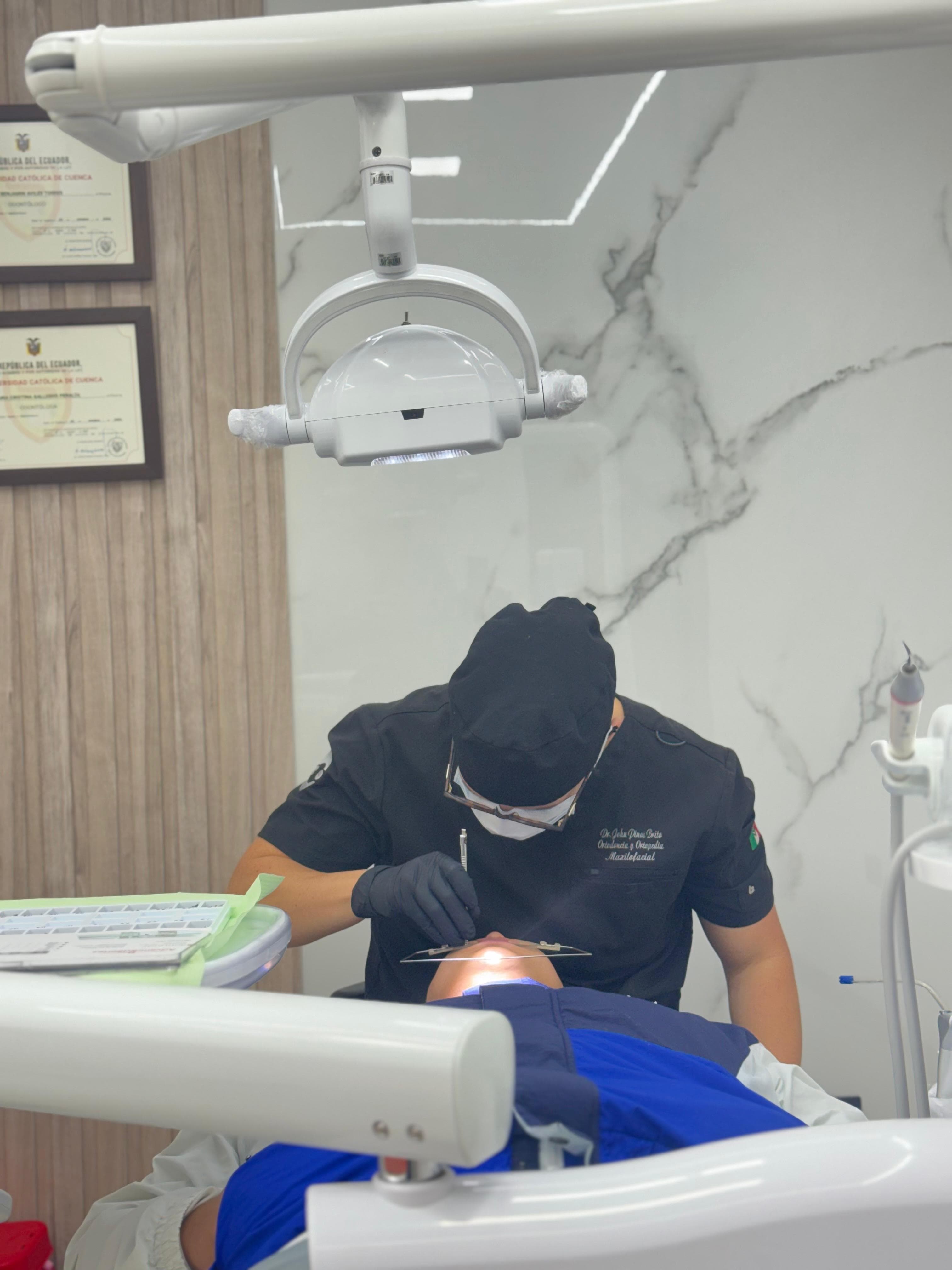 Dentist in black scrubs performs a procedure on a patient under a bright dental light.